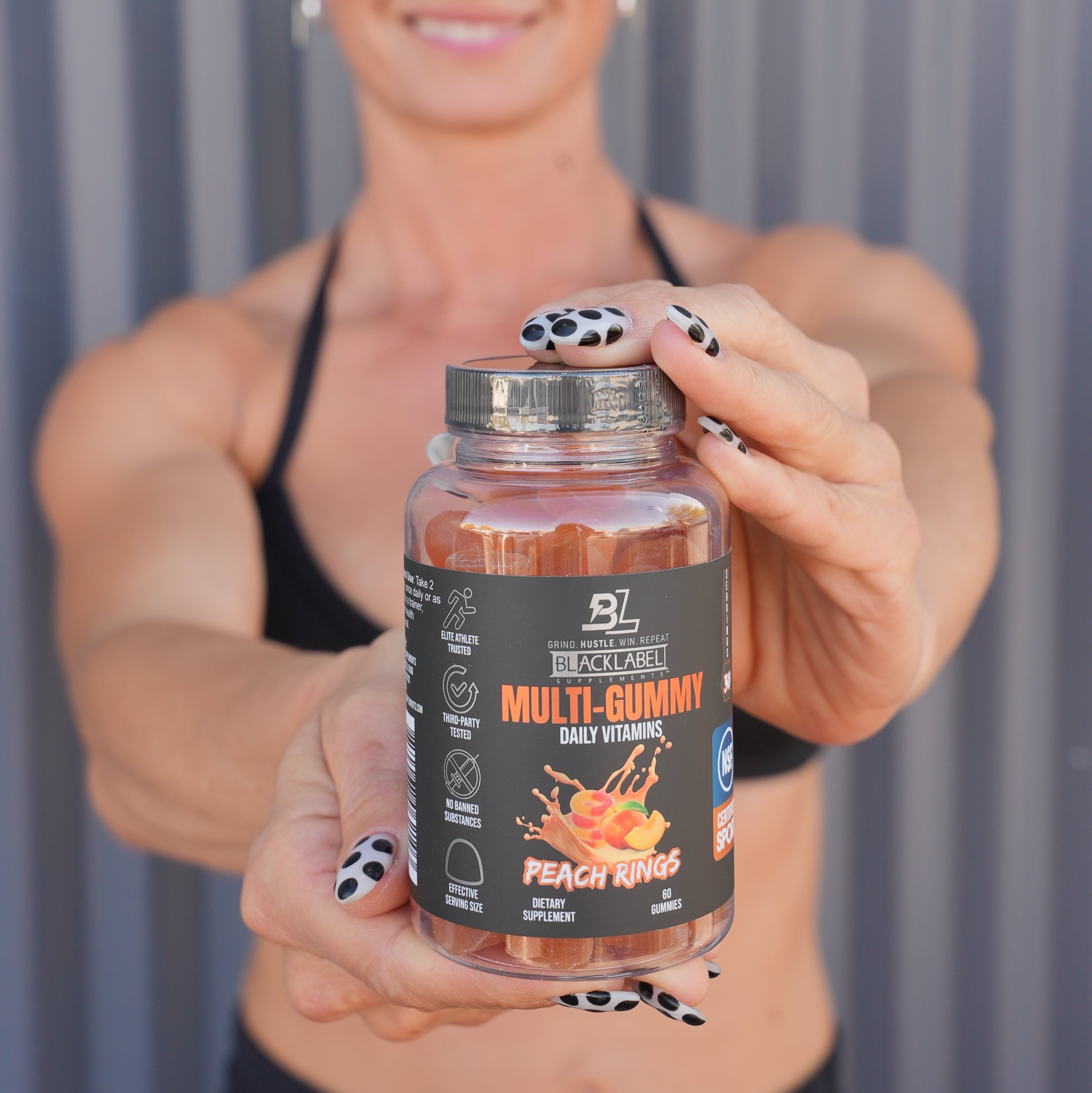 BlackLabel Supplements Multi Gummy Peach Rings in a clear jar, held by a woman with black and white polka dot nail polish, against a gray background.