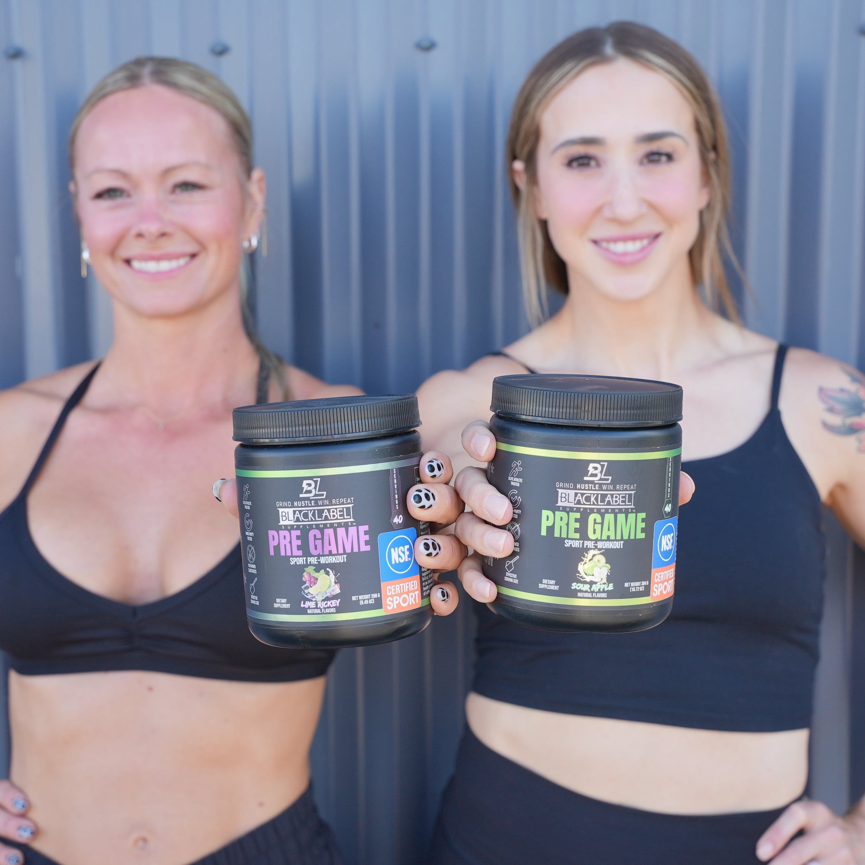 Two women in athletic wear holding containers of BLACKLABEL Supplements PRE GAME powder, standing in front of a metallic background.