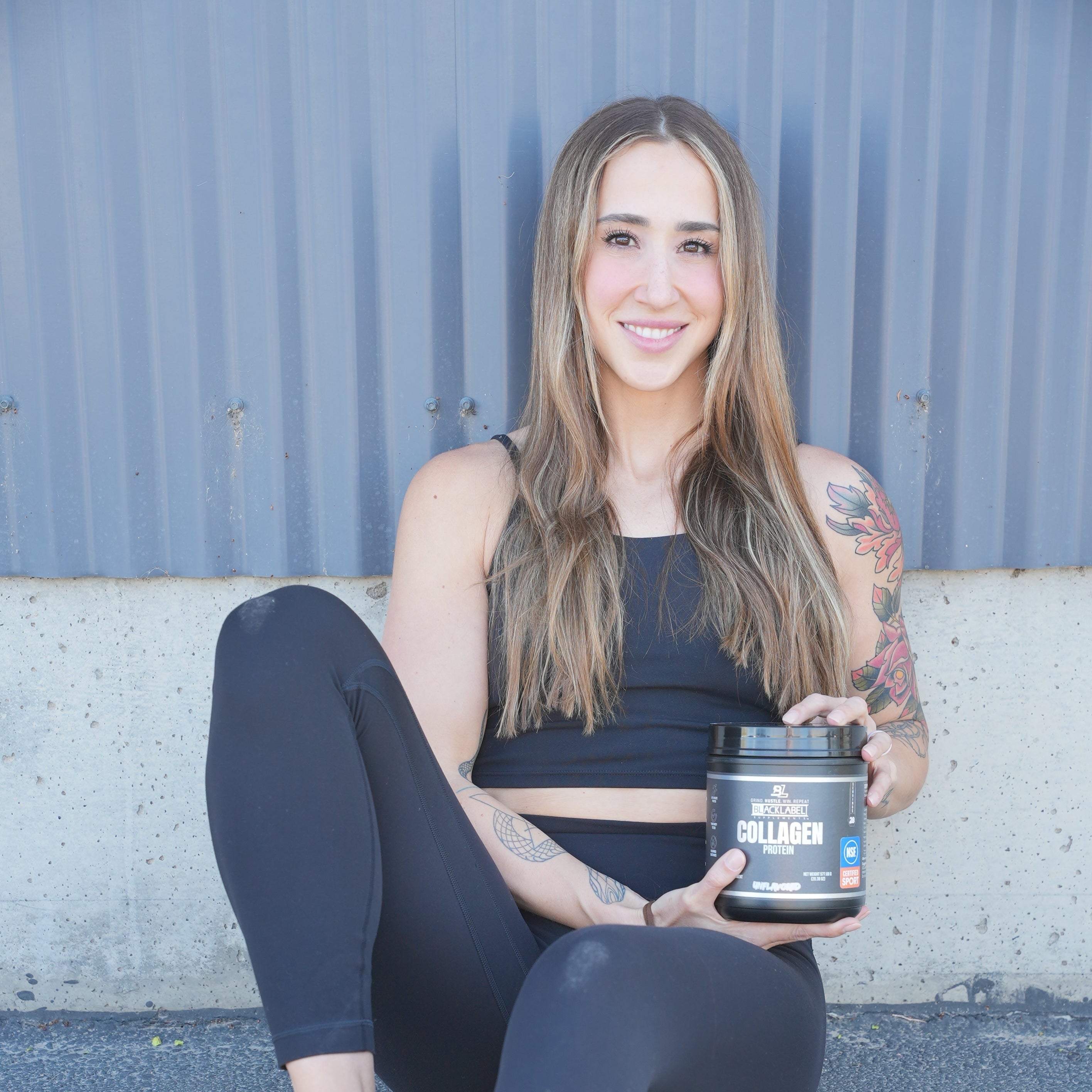 Young woman sitting outdoors holding a black container of BLACKLABEL Supplements Collagen Protein powder, wearing black athletic clothing.